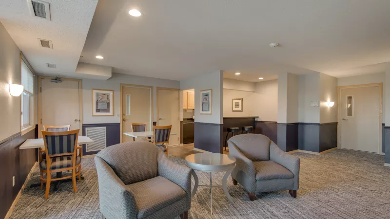 Inviting common area with two grey armchairs, a small round table, and a bar counter. Light walls, dark wainscoting, patterned carpet.
