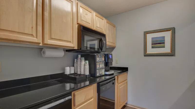 Small break room kitchen with light wood cabinets, black counters, a coffee machine, microwave, and framed art on a gray wall.
