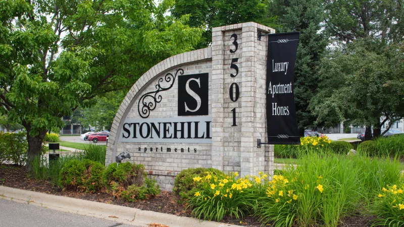 Stonehill Apartments entry sign with '3501', a banner for Luxury Apartment Homes, set amidst green landscaping and yellow flowers.