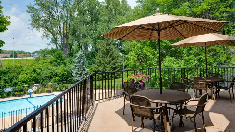 Outdoor patio with dining tables and umbrellas next to a blue swimming pool, surrounded by green trees.