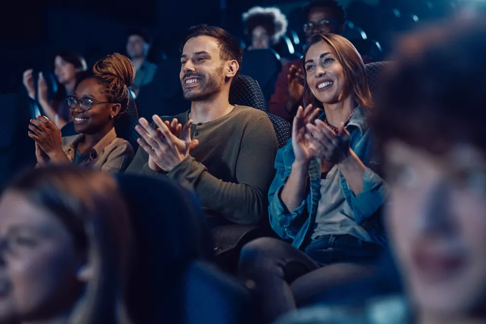 Diverse audience smiling and clapping in a dark movie theater, illuminated by screen light.