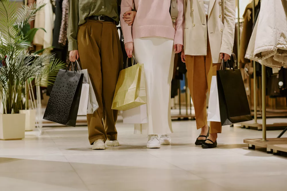 Three people walk through a clothing store, carrying shopping bags. One wears green and brown, another pink and white, a third cream.